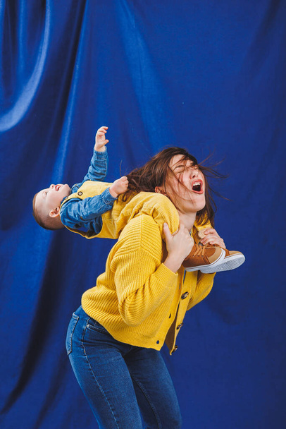 A young mother plays with her child on a blue background. Family relationship with the child. Raising a child, playing with a child - Photo, Image