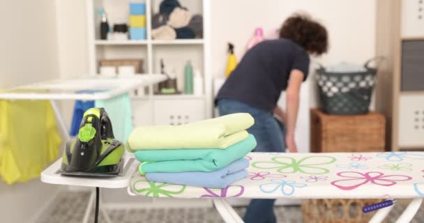 Adolescente trabajador con camisa azul, planchas en la tabla de planchar, se encarga de la ropa, realiza las tareas domésticas en el cuarto de lavado - Metraje, vídeo