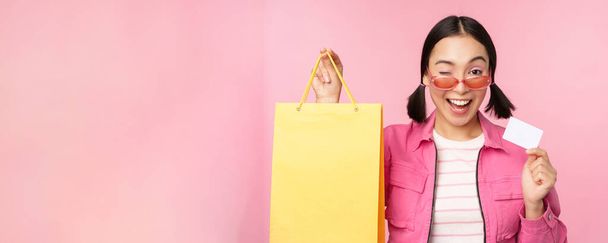 Heureuse jeune femme asiatique montrant carte de crédit pour faire du shopping, tenant sac, achat en vente, aller à la boutique, magasin, debout sur fond rose - Photo, image