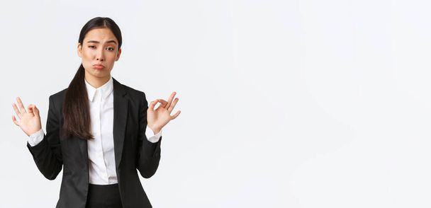 Pleased sassy asian businesswoman in black suit show not bad gesture, nod in approval and make okay gesture, very good work, praising coworker, standing satisfied over white background - Photo, Image