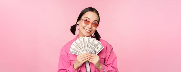Красива корейська жінка в сонцезахисних окулярах, показує долари, готівку, усміхнені задоволені, концепція швидких кредитів, мікрокредитування та оплати, стоїть на рожевому тлі
 - Фото, зображення