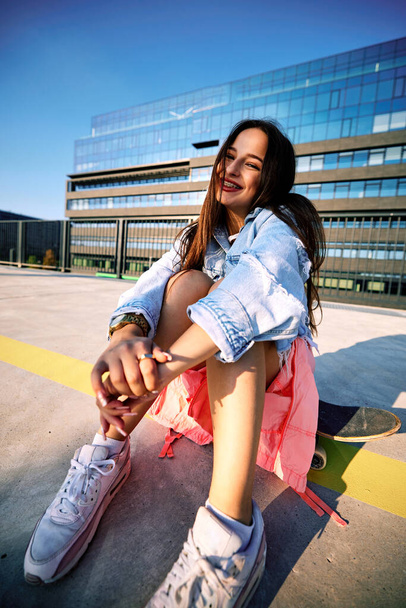 Uma adolescente skatista senta-se na estação de trem no skate e sorri para a câmera. - Foto, Imagem