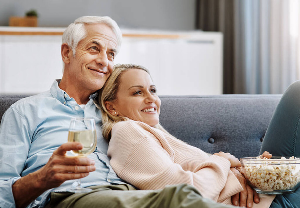Thuis veranderen in hun eigen bioscoop. Shot van een gelukkig volwassen paar ontspannen samen met wijn en popcorn op de bank thuis. - Foto, afbeelding