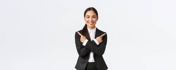 Μια όμορφη ασιάτισσα πωλήτρια προτείνει δύο επιλογές. Ελκυστική επιχειρηματίας με μαύρο κοστούμι δείχνει τα δάχτυλα πλάγια, έχουν λίγες παραλλαγές, να βοηθήσει να κάνουν την απόφαση, στέκεται λευκό φόντο - Φωτογραφία, εικόνα