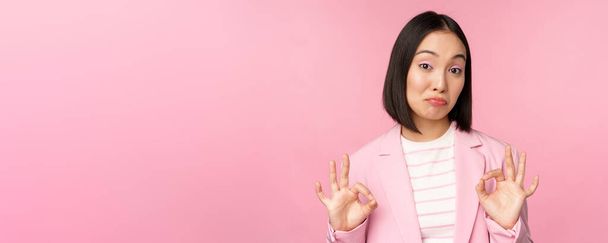 Неплохо. Впечатленная деловая женщина, азиатская офисная леди, показывающая в одобрении знак и кивок, стоящая на розовом фоне - Фото, изображение