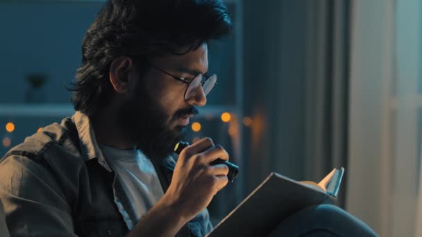 Árabe indio adulto hombre con barba usando gafas sentado en casa noche tarde oscuridad apagón lectura espiritual libro de texto con linterna casa escolaridad estudiar aprendizaje preparación para los exámenes - Imágenes, Vídeo
