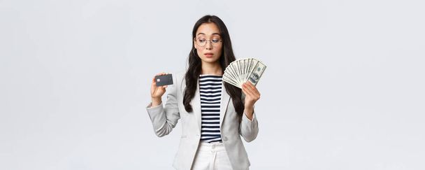 Business, finance and employment, entrepreneur and money concept. Indecisive cute asian office lady thinking about putting cash on deposit, looking curious at credit card, white background - Photo, Image