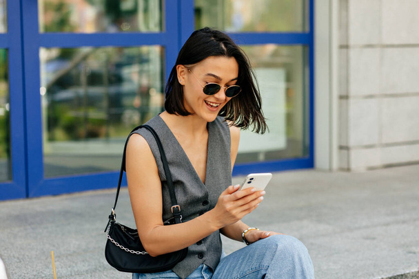 Pretty charming short-haired lady with wonderful smile is laughing outdoor with smartphone. Stylish woman in sunglasses and vest is sitting on stairs and scrolling smartphone - Foto, Imagem