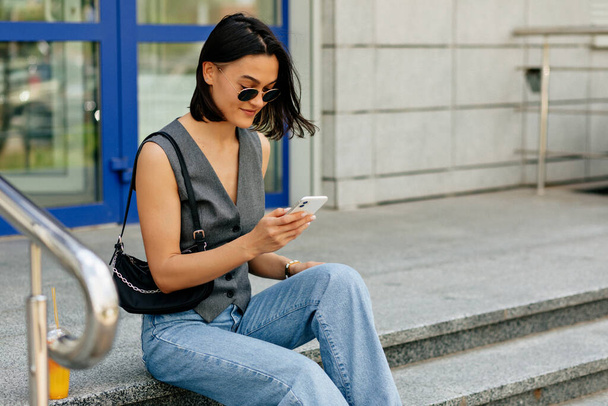 Stylowa kobieta w szarej kamizelce i dżinsach z torbą wysyła wiadomość przez telefon. Ładna pani uśmiecha się szczerze i siedzi na miejskich schodach.  - Zdjęcie, obraz