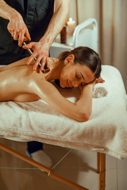 Happy female client getting back massage with hollow bamboo canes at spa salon - Photo, Image