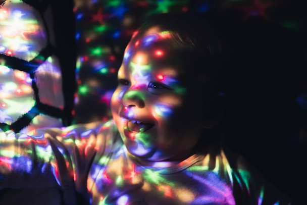 Caucasian toddler laughing while laying down on bed and looking at his brand-new lamp which is a colorful stars projector. - Photo, image
