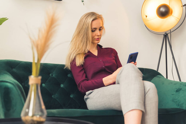 Σύγχρονη έννοια δραστηριοτήτων αναψυχής. Νεαρή καυκάσια ξανθιά γυναίκα εθισμένη στα μέσα κοινωνικής δικτύωσης κάθεται στον καναπέ της και αγοράζει ρούχα στο διαδίκτυο. - Φωτογραφία, εικόνα