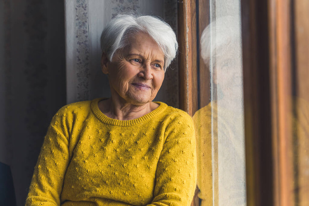 Innenaufnahme einer nachdenklichen älteren Rentnerin, die mit einem sanften Lächeln auf den Lippen durch das Fenster blickt. - Foto, Bild