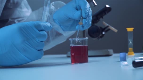 Chemisch laboratorium. Een onderzoeker in beschermende kleding pipetteert een monster van rode vloeistof uit een laboratoriumkolf. Close-up handen. - Video