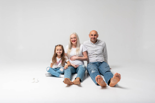 Glückliche dreiköpfige Familie auf dem Boden sitzend, heller Hintergrund. Lacht: Mutter, Vater und kleiner Junge. Mama ist schwanger. Gute Nachrichten, schwangere, nackte Füße für uns - Foto, Bild
