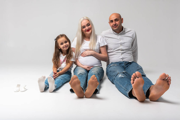 Χαρούμενη οικογένεια τριών ατόμων που κάθονται στο πάτωμα, με φόντο το φως. Γελάει: μητέρα, πατέρας και αγοράκι. Η μαμά είναι έγκυος. Καλά νέα, έγκυος, γυμνά πόδια μας - Φωτογραφία, εικόνα