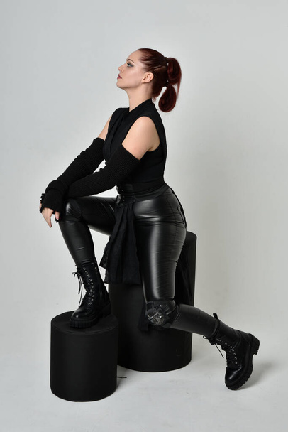 Volledig portret van mooie roodharige vrouwelijke model dragen zwarte futuristische scifi lederen mantel kostuum. Dynamisch zitten knielen pose op witte studio achtergrond. - Foto, afbeelding