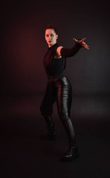 Ganztägiges Porträt des hübschen rothaarigen weiblichen Modells im schwarzen futuristischen Scifi-Ledermantel-Kostüm. Stehende Pose auf dunklem Studiohintergrund mit stimmungsvoller Beleuchtung am Schattenrand. - Foto, Bild