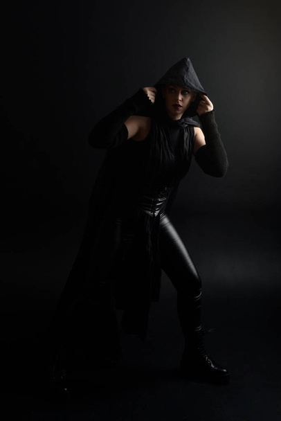 Full length portrait of pretty redhead female model wearing black futuristic scifi leather cloak costume. Standing pose on dark studio background with shadow rim  moody lighting. - Foto, afbeelding