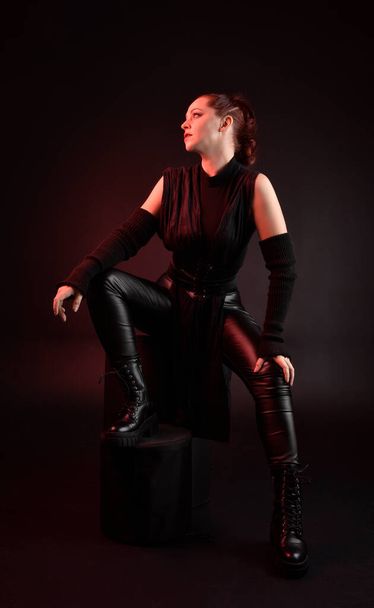 Siyah fütüristik deri pelerin giyen güzel kızıl saçlı kadın modelin tam boy portresi. Karanlık stüdyo arka planında oturan karamsar ışıklandırmalı.. - Fotoğraf, Görsel