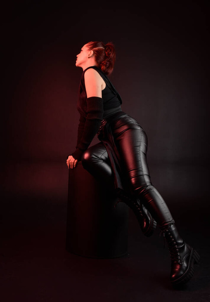 Portrait pleine longueur de jolie rousse modèle féminin portant noir futuriste manteau en cuir scifi costume. pose assise sur fond de studio sombre avec éclairage lunaire de bord d'ombre. - Photo, image