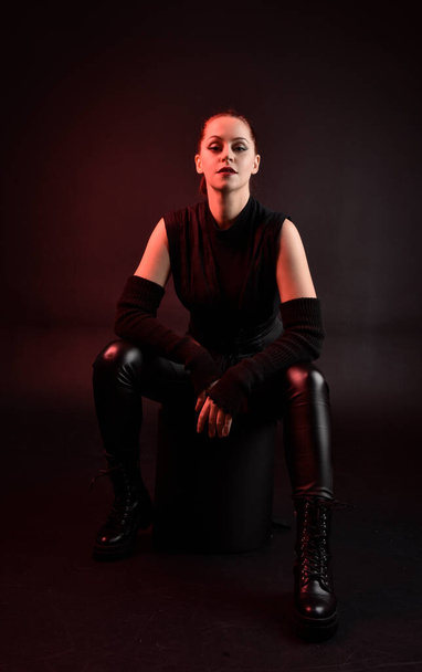 Ganztägiges Porträt des hübschen rothaarigen weiblichen Modells im schwarzen futuristischen Scifi-Ledermantel-Kostüm. Sitzpositionen auf dunklem Studiohintergrund mit stimmungsvoller Beleuchtung am Schattenrand. - Foto, Bild