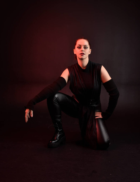 Full length portrait of pretty redhead female model wearing black futuristic scifi leather cloak costume. Sitting  pose on dark studio background with shadow rim  moody lighting. - Φωτογραφία, εικόνα