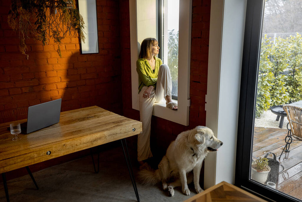 Frau sitzt auf der Fensterbank und schaut zu Hause mit ihrem weißen liebenswerten Hund aus dem Fenster - Foto, Bild