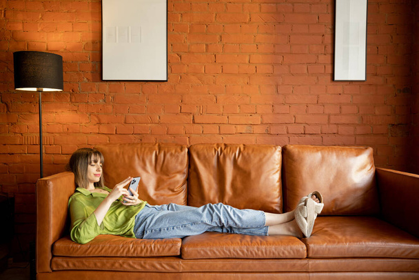 Woman using phone while relaxing on a leather couch at home - Foto, immagini
