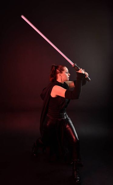 Full length portrait of pretty redhead female model wearing black futuristic scifi leather cloak costume. Standing pose  holding lightsaber on dark studio background with shadow moody lighting. - Photo, Image