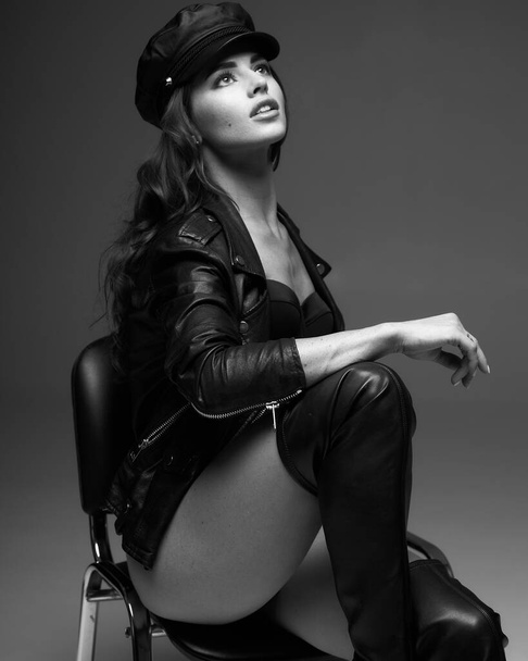 Portrait of young attractive woman model with brown long curly hair, leather jacket and hat, long legs with tan skin, slim body, sitting on soft gray background. The concept of a beautiful elegant figure, body positive, healthy lifestyle. - Фото, изображение