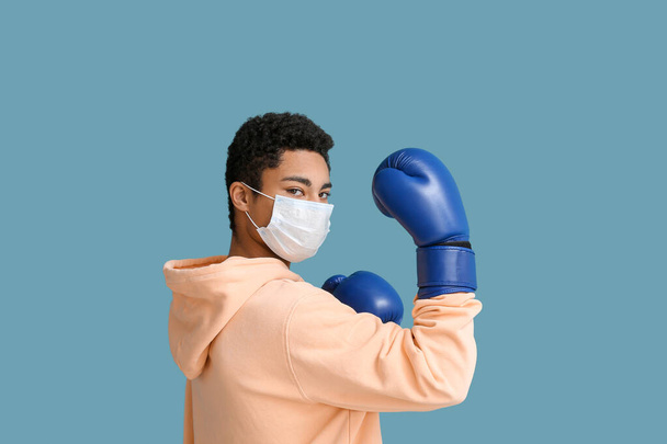 Nuori afroamerikkalainen mies lääketieteellisessä naamiossa nyrkkeilyhanskat sinisellä pohjalla. Koskemattomuuden käsite - Valokuva, kuva