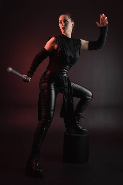 Full length portrait of pretty redhead female model wearing black futuristic scifi leather cloak costume. Standing pose  holding lightsaber on dark studio background with shadow moody lighting. - 写真・画像
