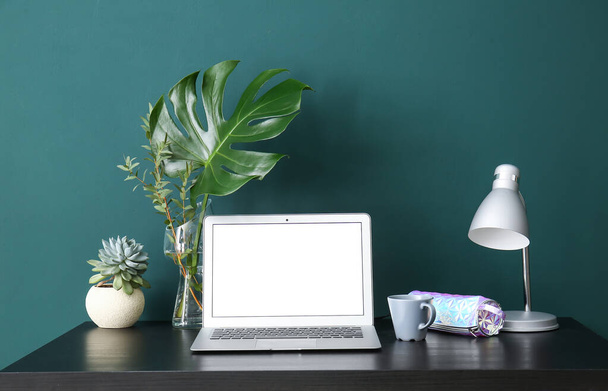 Local de trabalho com laptop, ramos de plantas em vaso, lâmpada e xícara perto da parede verde - Foto, Imagem