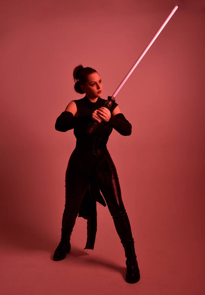 Full length portrait of pretty redhead female model wearing black futuristic scifi leather cloak costume. Standing pose  holding lightsaber on red studio background with coloured lighting - Foto, imagen