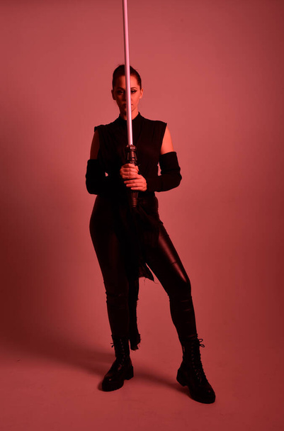 Full length portrait of pretty redhead female model wearing black futuristic scifi leather cloak costume. Standing pose  holding lightsaber on red studio background with coloured lighting - Фото, зображення
