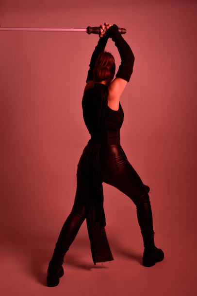 Full length portrait of pretty redhead female model wearing black futuristic scifi leather cloak costume. Standing pose  holding lightsaber on red studio background with coloured lighting - Foto, afbeelding