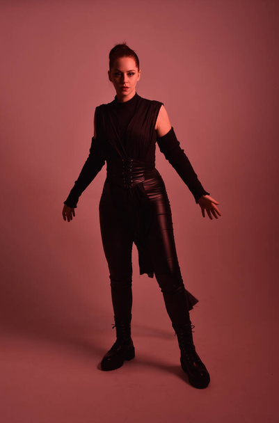 Full length portrait of pretty redhead female model wearing black futuristic scifi leather cloak costume. Standing pose  holding lightsaber on red studio background with coloured lighting - Zdjęcie, obraz