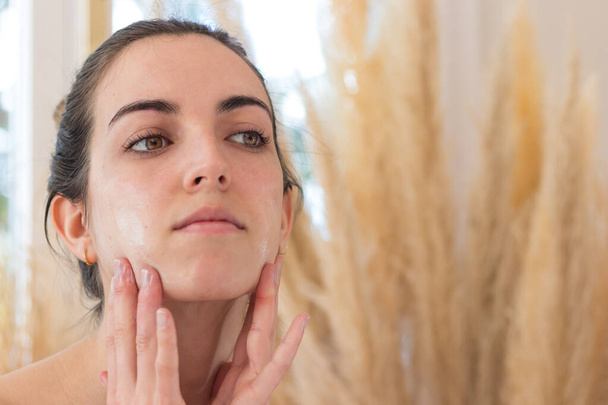 Woman looking to the mirror while applying moisturizer to her flawless skin - Foto, Bild