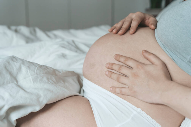 Ciężarna kobieta głaska się po brzuchu z dzieckiem przed porodem, relaksując się w słonecznej, jasnej sypialni leżącej na łóżku. Zdrowie kobiet, ciąża, poczęcie, koncepcja porodu. - Zdjęcie, obraz