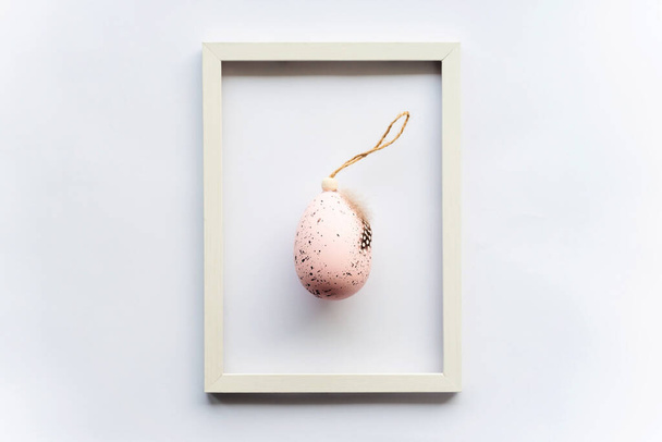 Πασχαλινό αυγό σε ροζ χρώμα με φτερό πουλιού σε λευκό φόντο με χώρο αντιγραφής για κείμενο σε πλαίσιο. Ελάχιστη έννοια. Θέα από ψηλά. Κάρτα Πάσχα, μαλακή επιλεκτική εστίαση. - Φωτογραφία, εικόνα