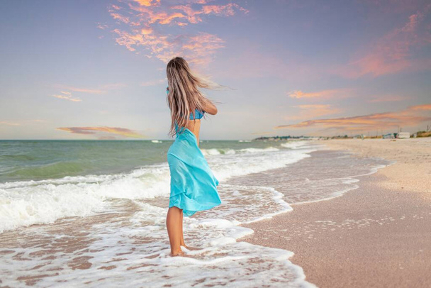 A young slender tanned girl with blonde hair in a gentle bluish swimsuit and a bright light blue shawl, walks along the beach near the edge of the sea water enjoying the warm summer - Foto, Bild