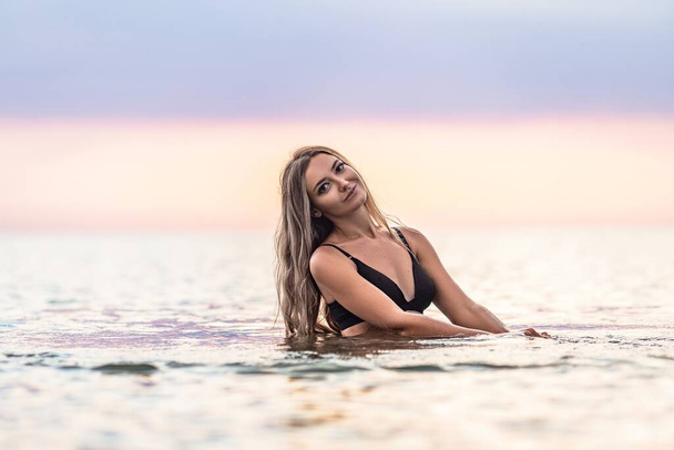 A slender tanned young girl with blond lush hair in a black swimsuit, splashes to the sides while sitting in a shallow estuary against the background of a bright warm sunset - Valokuva, kuva