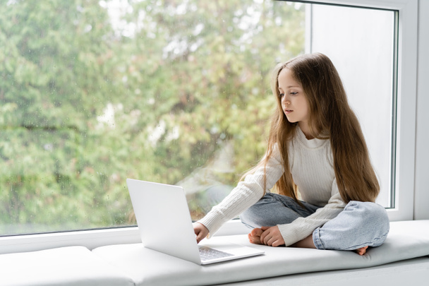 κορίτσι κάθεται στο περβάζι με σταυρωμένα πόδια και τη χρήση φορητού υπολογιστή κατά τη διάρκεια του online μαθήματος - Φωτογραφία, εικόνα