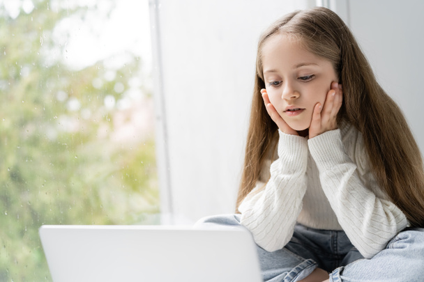 menina focada de mãos dadas perto do rosto enquanto olha para laptop perto da janela - Foto, Imagem