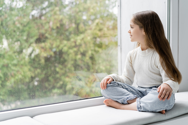 blootsvoets meisje in jeans zittend op vensterbank met gekruiste benen en kijkend door glas met regendruppels - Foto, afbeelding