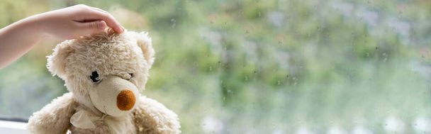vista parcial de la mano del niño cerca del oso de peluche y ventana borrosa con gotas de lluvia, pancarta - Foto, Imagen