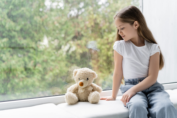 girl in jeans sitting on windowsill near teddy bear and glass with raindrops - Photo, Image