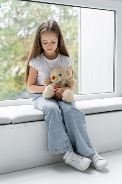 full length view of girl holding teddy bear while sitting on windowsill - Foto, Bild