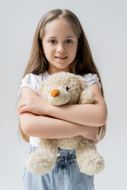 zufriedenes Mädchen umarmt Teddybär, während es isoliert auf grau in die Kamera schaut - Foto, Bild